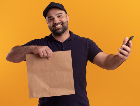微笑微笑着的中年送货员 穿着制服 戴着帽子 手里拿着纸食品包 电话被隔离在黄色的墙上人拿着人