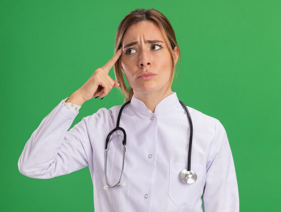 脸想着看着身边年轻的女医生穿着医用长袍 用听诊器把手指放在隔离在绿墙上的太阳穴上人人感情