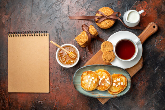 螺旋形新鲜煎饼俯视图一杯红茶放在木制砧板上蜂蜜饼干牛奶和螺旋笔记本堆在黑暗的表面上顶部视图午餐