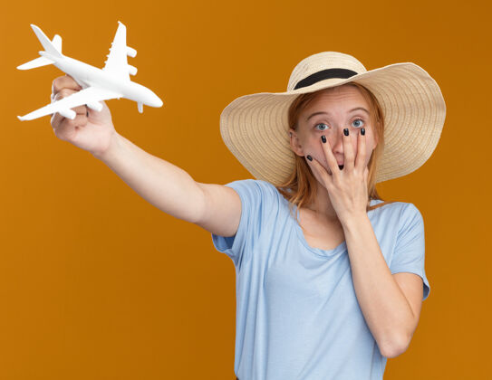 橙色震惊的年轻红发姜女雀斑戴沙滩帽把手放在嘴上 并持有模型飞机模型手飞机