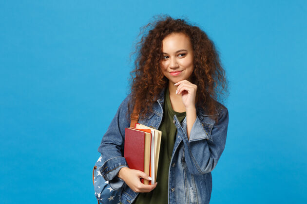 学习穿着牛仔服 背着书包的年轻女学生把书孤立地放在蓝色的墙上教学校园人