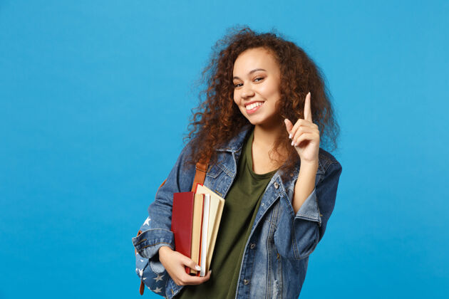 阅读穿着牛仔服 背着书包的年轻女学生把书孤立地放在蓝色的墙上学生信息教学