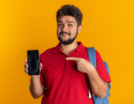 年轻年轻的留着胡子的学生 穿着红色马球衫 背着背包 拿着智能手机 食指指着智能手机 微笑着 自信 快乐 积极地站在橙色的墙上手指站立正面