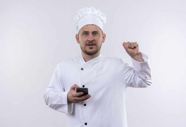 烹饪自信的年轻帅哥厨师身着厨师制服手持手机举起拳头孤立在白墙上自信拳头持枪