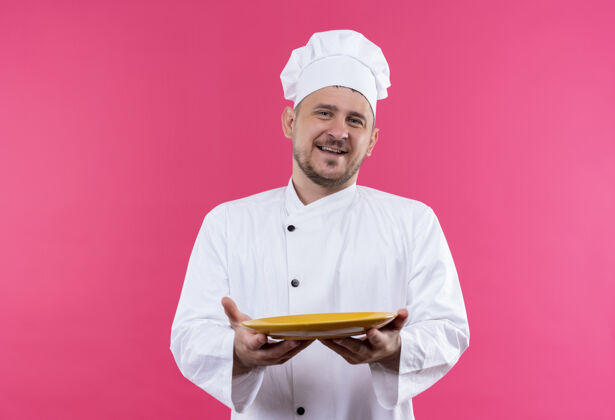 舒展快乐的年轻帅哥厨师穿着厨师制服 把空盘子摊在粉红色的墙上烹饪帅气盘子