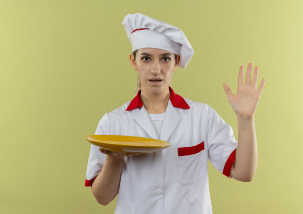 印象年轻漂亮的厨师穿着厨师制服 手里拿着空盘子 举起一只孤立在绿墙上的手漂亮手烹饪