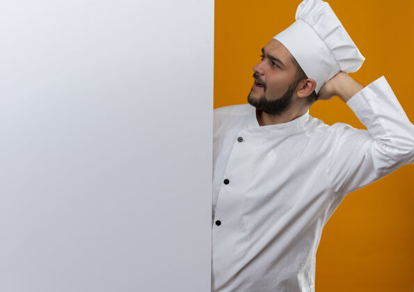 手困惑的年轻男厨师穿着厨师制服站在后面 看着白色的墙壁 手放在脑后 隔离在橙色的墙壁上 有复制空间男厨师年轻