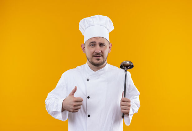 帅气自信的年轻帅哥厨师 身着厨师制服 拿着勺子 在隔离的橙色墙壁上竖起大拇指 留着复印空间制服拇指抱