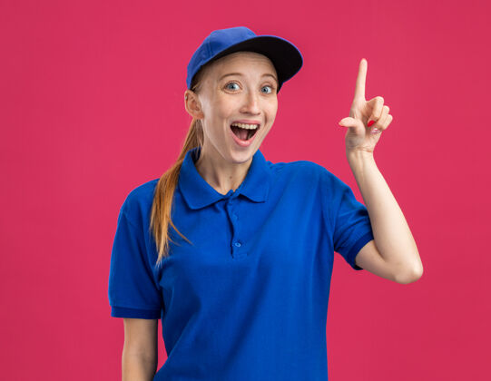 帽子身穿蓝色制服 戴着帽子的年轻送货员惊讶而高兴地把有新想法的食指放在粉红色的墙上目录女孩年轻