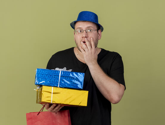手震惊的成年斯拉夫男子戴着眼镜 戴着蓝色派对帽 手放在脸上 手里拿着礼品盒和纸购物袋眼镜礼物持有