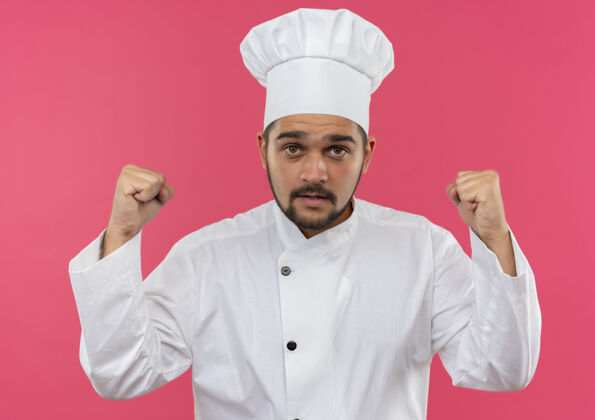 提高令人印象深刻的年轻男厨师在厨师制服举起拳头和孤立的粉红色墙上厨师烹饪男性