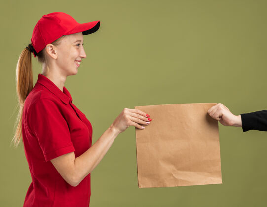 女孩年轻的送货员穿着红色制服 戴着帽子 手里拿着一个纸包 在绿色的墙壁上自信地微笑着送给顾客给予送货年轻