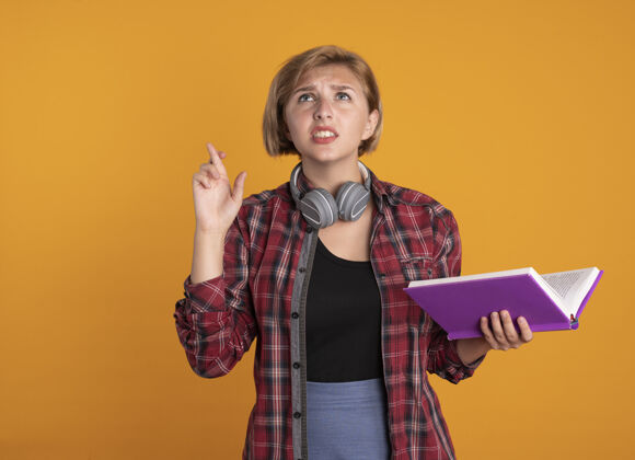 耳机焦急的年轻斯拉夫女学生戴着耳机背着书包 手指交叉 手里拿着书年轻穿学生