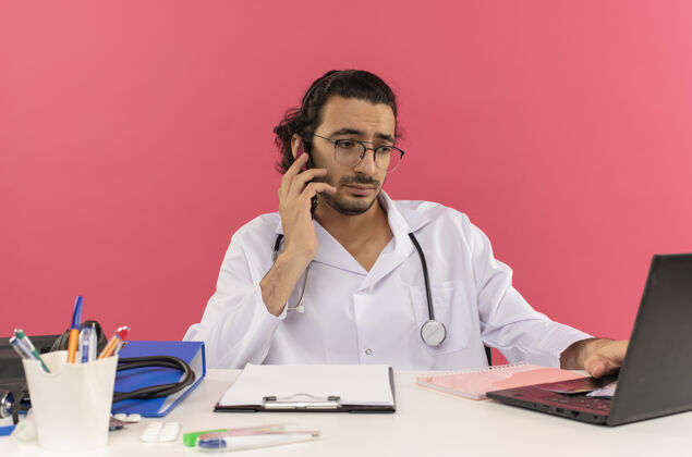 空间悲哀的年轻男医生 戴着医用眼镜 穿着医用长袍 听诊器坐在桌子旁粉色医生悲伤
