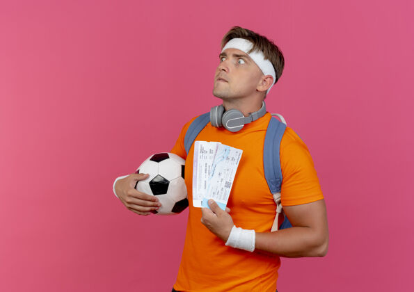 男人给人印象深刻的年轻英俊的运动型男人戴着头带和腕带 脖子上背着一个戴着耳机的包 手里拿着机票和足球 看上去笔直飞机包复制