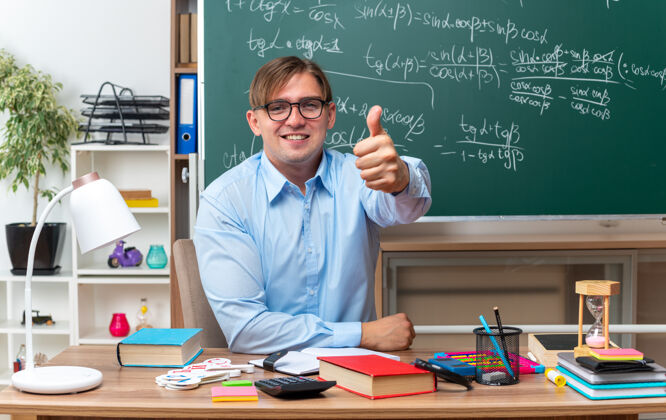 展示年轻的男老师戴着眼镜 面带微笑 自信地竖起大拇指坐在课桌旁 教室的黑板前放着书和笔记数学坐着年轻