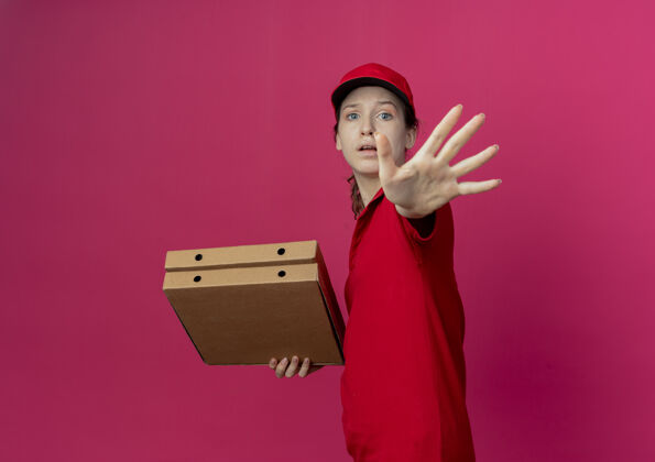 深红色穿着红色制服 戴着帽子的年轻漂亮送货女孩站在侧视图中 手里拿着披萨包 伸出手示意不年轻制服红色