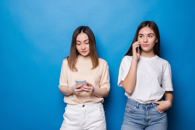 快乐年轻女孩在讲电话 另一个女孩在蓝色的墙上用电话使用手机时尚表情
