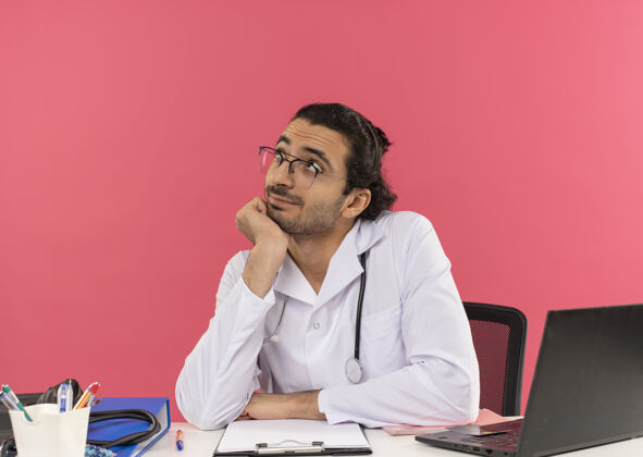 手年轻的男医生戴着医用眼镜 穿着医用长袍 带着听诊器坐在办公桌旁 高兴地看着我副本穿坐着