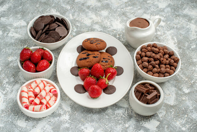 糖果白色椭圆形盘子上的巧克力饼干草莓和圆形巧克力 灰白色背景上的碗里有糖果草莓巧克力麦片和可可覆盆子巧克力草莓
