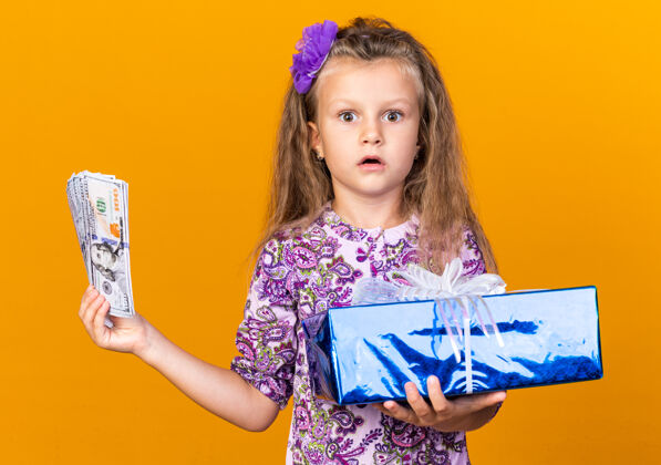 女孩惊讶的小金发女孩拿着礼品盒和钱孤立在橙色墙上的复制空间持有小钱