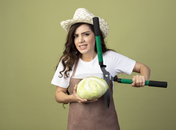 帽子穿着制服的年轻女园丁戴着园艺帽 把卷心菜和园艺剪孤立地放在橄榄绿的墙上人姿势不愉快