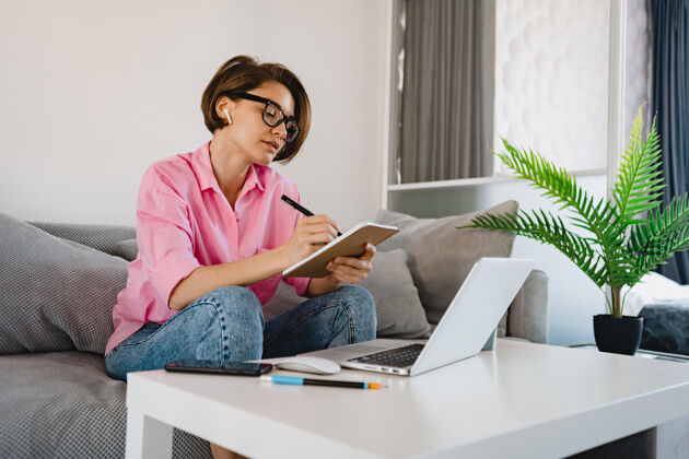 设备穿着粉色衬衫的忙碌严肃的女人坐在沙发上专心做笔记 在桌子上付帐单 在家里用笔记本电脑上网无线自由职业者成人