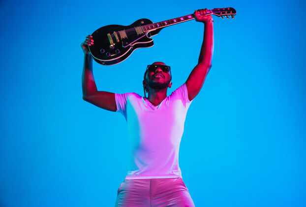 艺术家年轻的非洲裔美国音乐家在霓虹灯下像摇滚明星一样在蓝色的墙上弹吉他男人球员成人