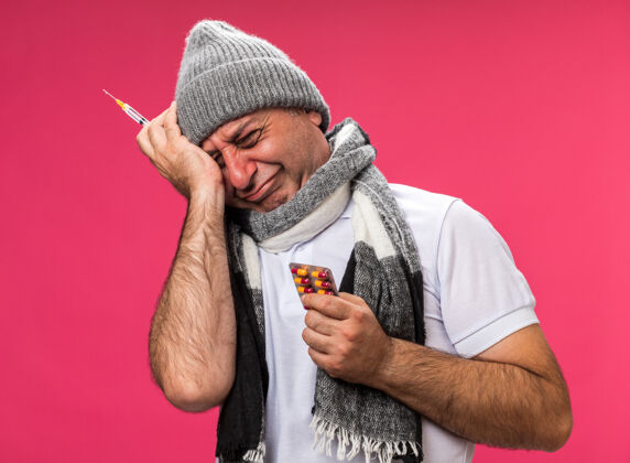 孤立悲伤的成年病高加索男子脖子上围着围巾戴着冬季帽子拿着注射器和药物水泡包隔离在粉红色墙上的复制空间水泡悲伤围巾