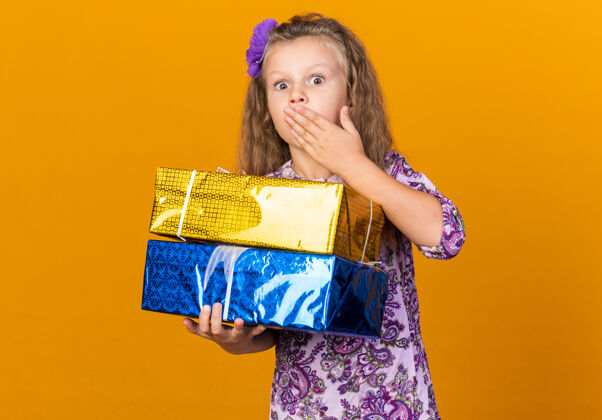 盒子惊讶的小金发女孩把她的手放在她的嘴上 拿着礼品盒隔离在橙色墙上的复制空间金发小拿着
