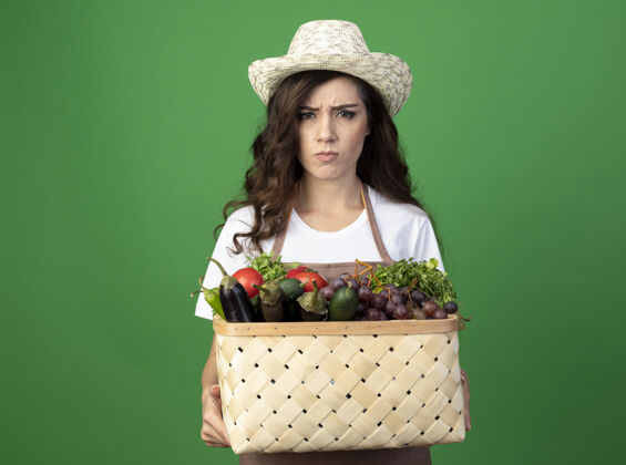 年轻人穿着制服的年轻女园丁戴着园艺帽 把菜篮子孤立地放在绿色的墙上不愉快人姿势