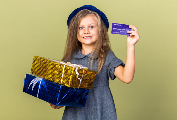 派对惊讶的金发小女孩 戴着蓝色派对帽 拿着礼品盒和信用卡 隔离在橄榄绿的墙上 留着复印空间生日女孩小
