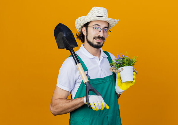 花园年轻的留着胡须的园丁 穿着连体衣 戴着帽子 拿着铲子和盆栽植物 站在橙色的墙上 面带微笑地看着前方感觉微笑衣服