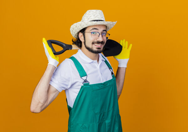 连身衣年轻的留着胡须的园丁穿着连体衣 戴着帽子 拿着铲子 站在橘色的墙上 兴高采烈地微笑着看着前面感觉胡须年轻人