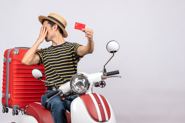 帽子一个年轻人戴着草帽 手拿信用卡 把脸藏在摩托车上前面信贷摩托车