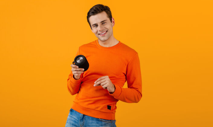 模特穿着橙色毛衣的时髦微笑的年轻人拿着无线扬声器快乐地听音乐享受橙色的乐趣积极年轻便携