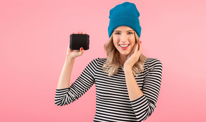 模特年轻女子手持无线扬声器听音乐穿着条纹衬衫和蓝色帽子微笑着摆出粉红色的姿势乐趣音频享受