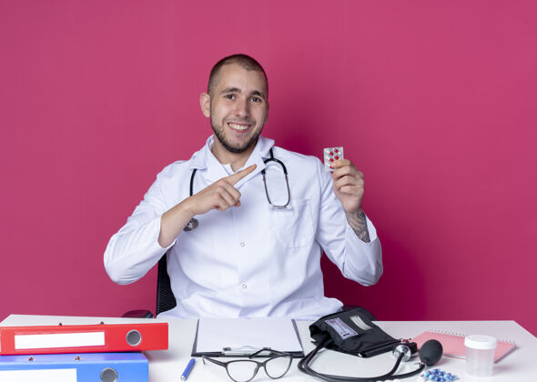 医学面带微笑的年轻男医生穿着医用长袍 听诊器坐在办公桌旁 手里拿着工作工具 指着隔离在粉红色墙上的一包胶囊长袍表情人