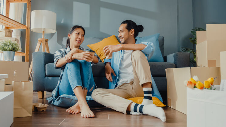 人快乐的亚洲年轻夫妇男女坐在新家里喝咖啡和聊天用纸箱包装箱存放女性休闲房间