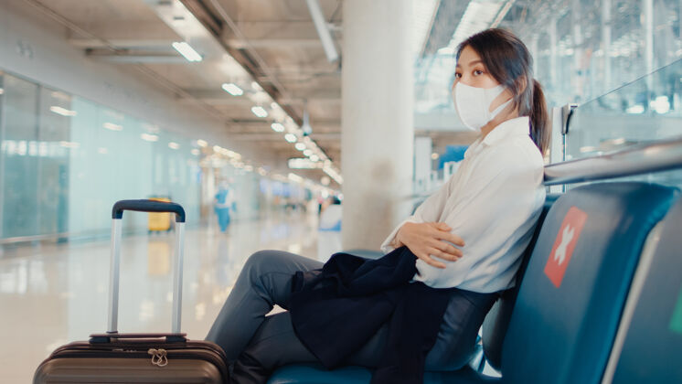 航班亚洲商务小姐拿着行李坐在长凳上等待和寻找伙伴的航班在机场乘客终端面子