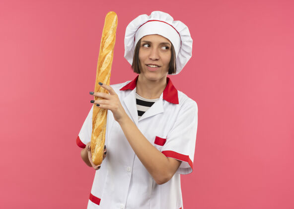厨师面带微笑的年轻女厨师身着厨师制服拿着面包棒看着隔离在粉红色墙上的一面姿势表情制服