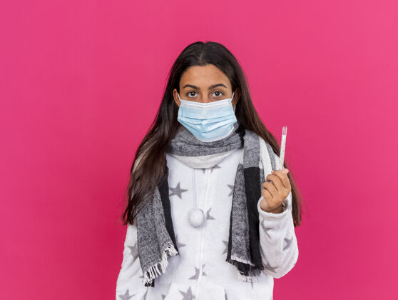 穿年轻的生病女孩戴着医用面罩 戴着围巾 拿着隔离在粉色上的温度计持有体温计年轻