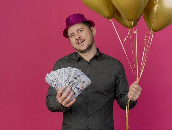 年轻人高兴的年轻人戴着粉红色的帽子拿着现金和气球孤立在粉红色的聚会戴着气球粉色