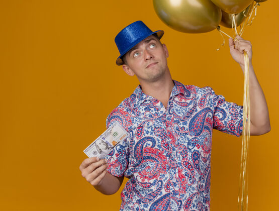 抱令人印象深刻的年轻人戴着蓝色帽子拿着气球现金孤立在橙色壁板帽子气球
