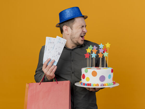 聚会快乐的年轻人闭着眼睛戴着蓝色帽子 拿着礼品袋和蛋糕 票子被隔离在橙色上蛋糕举行穿