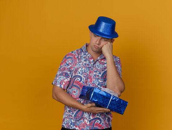思考一个体贴的年轻人 戴着蓝色帽子 拿着礼盒 手放在橘子色的脸颊上 看着下面蓝色橙色拿着
