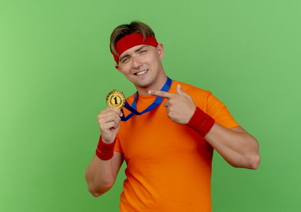 指着高兴的年轻英俊的运动男子戴着头带和腕带 脖子上拿着奖牌 指着隔离在绿色墙上的奖牌脖子帅气市民