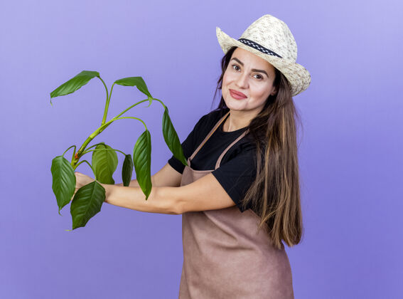 园艺高兴美丽的园丁女孩穿着制服 戴着园艺帽 在蓝色隔离的一边伸出植物请蓝色花园