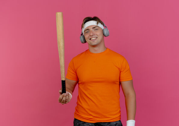 微笑面带微笑的年轻英俊的运动型男子戴着头带和腕带 头戴耳机拿着棒球棒孤立地站在粉红色的墙上运动人姿势
