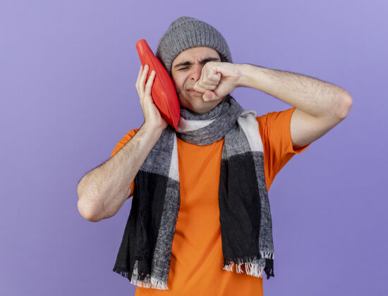 湿巾年轻的病夫戴着冬天的帽子戴着围巾把热水袋放在脸颊上擦脸用手隔离在紫色上脸颊帽子眼睛